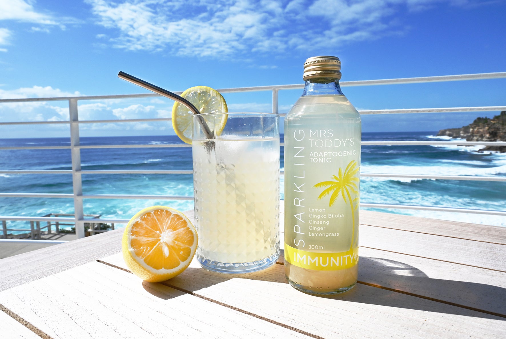 Mrs. Toddy’s Sparkling Immunity Adaptogenic Tonic bottle sits on a sunlit wooden table beside a chilled glass with a lemon slice and metal straw, overlooking a bright blue ocean and clear sky—perfectly capturing a refreshing coastal wellness vibe.